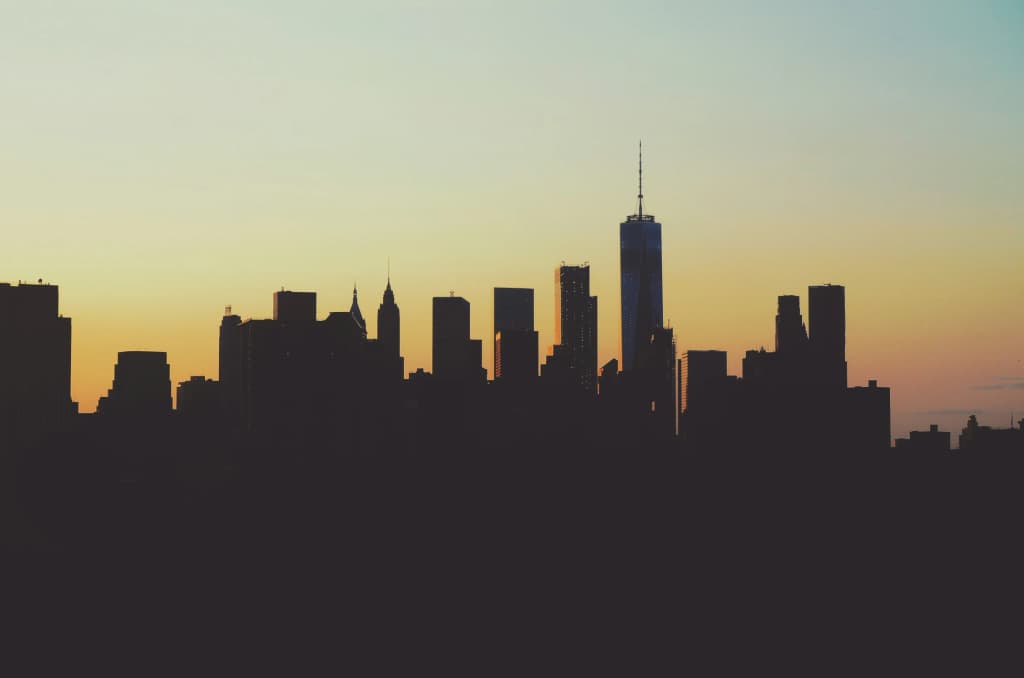 Silhouetted Lower Manhattan skyline with One World Trade Center against a yellow and teal sunset sky