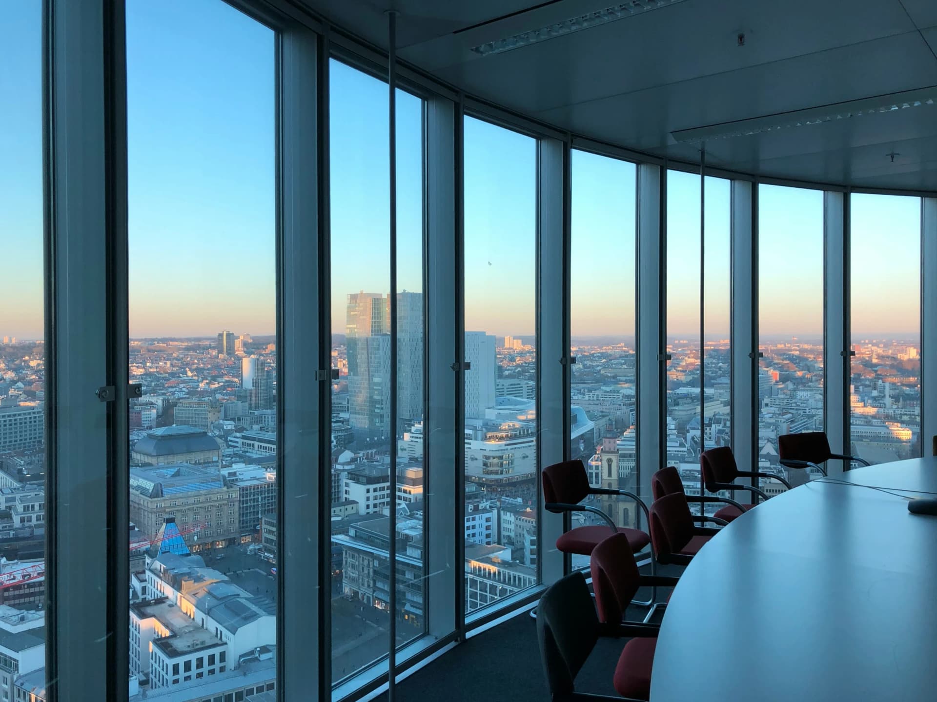 Empty conference room with red chairs around a curved table, behind floor-to-ceiling windows overlooking a city skyline at sunset
